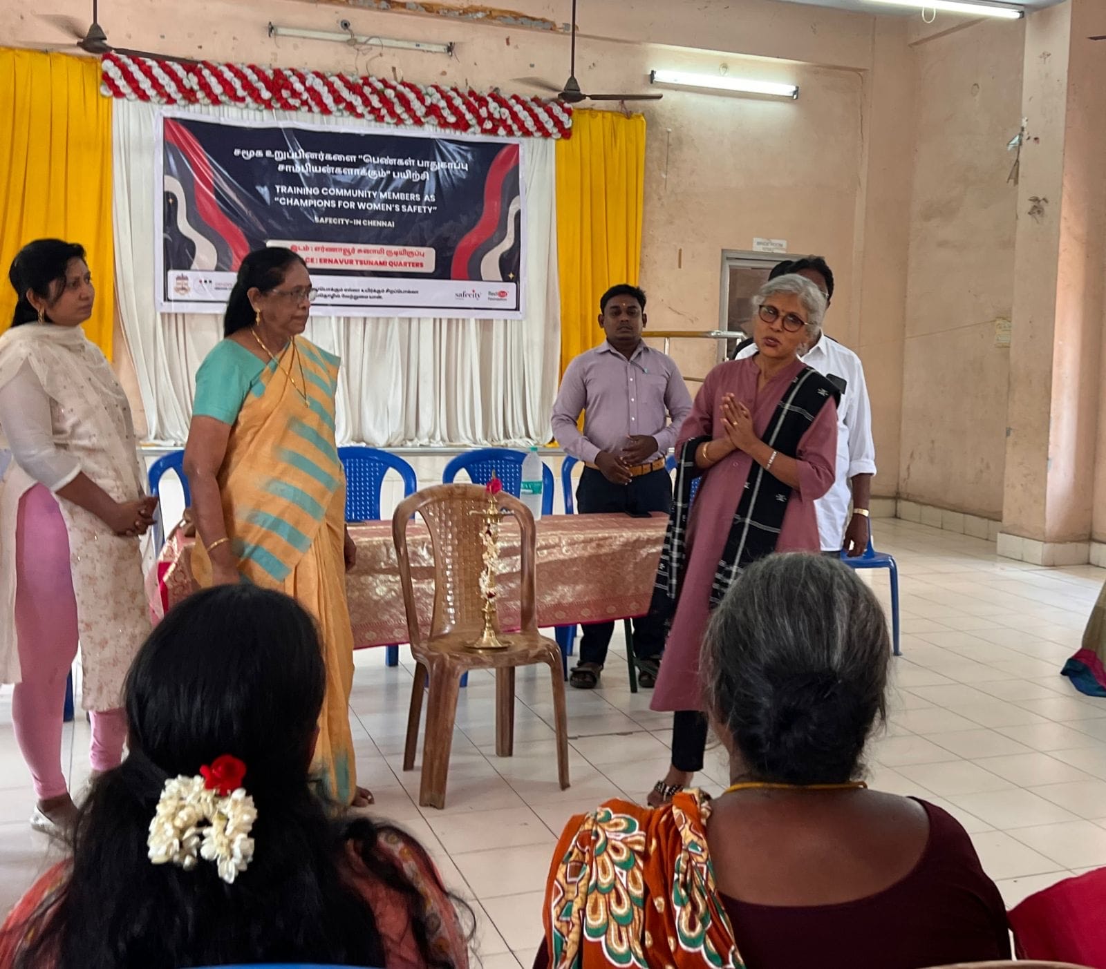 An image of people engaged in a workshop on safety training for women. The location is a room with some people seated on chairs facing a few others who are standing in front and speaking to the gathered. 