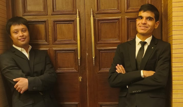 Image of Mihaan and Vir. Both are wearing black and white suits and leaning against a beautifully carved door.
