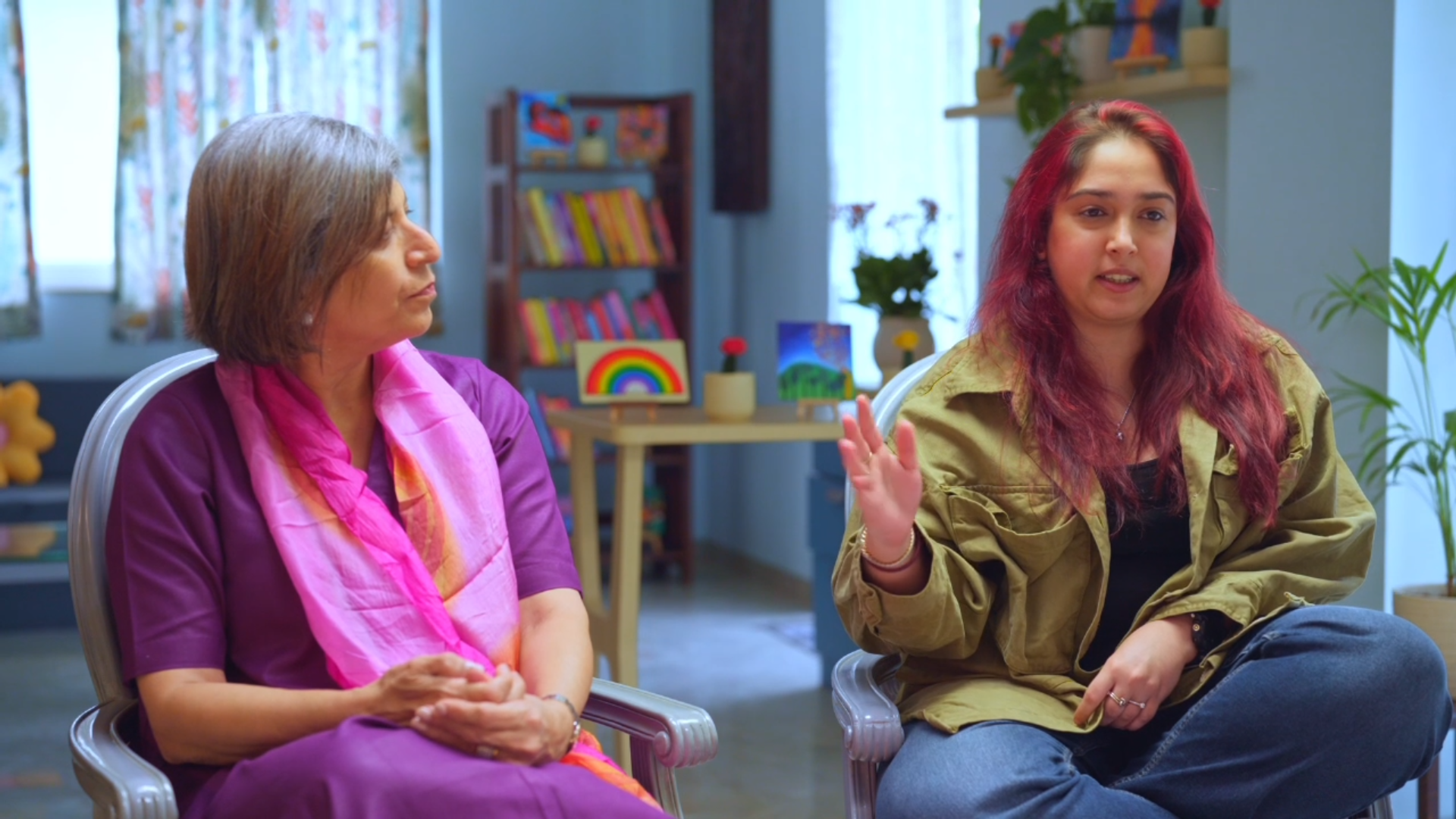 A still from show - Parenting Aaj Kal with Ira Khan with Dr. Parven. Two women sit indoors, engaged in conversation. Dr. Parven on the left wears a purple outfit with a pink scarf, while Ira Khan on the right, with red hair, wears a green jacket and gestures with her hand.