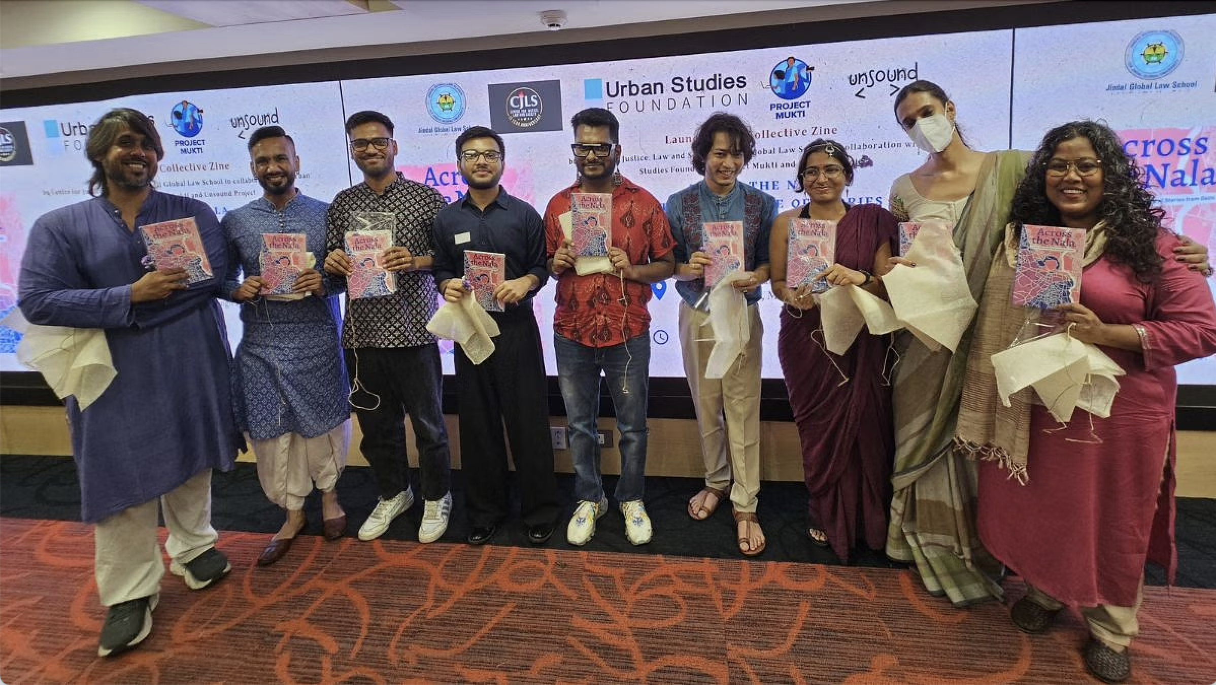 The image shows a group of ten people standing in a line indoors, each holding a colorful book titled "Across the Nala." They are positioned in front of a large digital screen displaying several logos and event information. The individuals are dressed in a variety of outfits, ranging from casual to traditional attire, and each person is smiling towards the camera. The floor has a patterned red carpet. Some individuals are holding white cloth materials, giving the impression of a celebratory or formal event.  Alt-text:  A group of ten people holding books titled "Across the Nala" in front of a digital screen with event information.